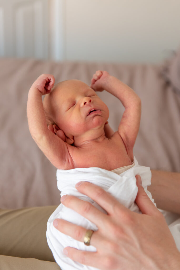 Newborn baby stretching arms wide while dad holds baby during relaxed lifestyle photo session at home in St. George, Utah