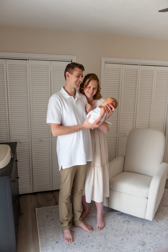 Full body photo of mom and dad gazing lovingly at their newborn during lifestyle session in their St. George home